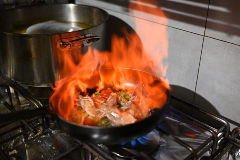 Τηγάνι με φαγητό που φλέγεται πάνω σε εστία κουζίνας, με κατσαρόλα στο πλάι.